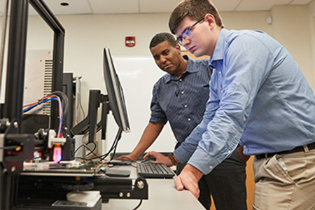 Two male students lean over looking at a computer in the Computers and Technology program