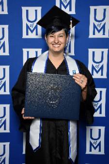 angela_g April holds her diploma while smiling in her black graduation gown and cap. UofM logo wall background behind her.