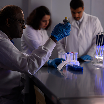Three researchers in white lab coats are working in a lab with pipettes and test tubes