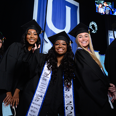Three female grads in commencement regalia and caps smiling before walking across the stage to graduate