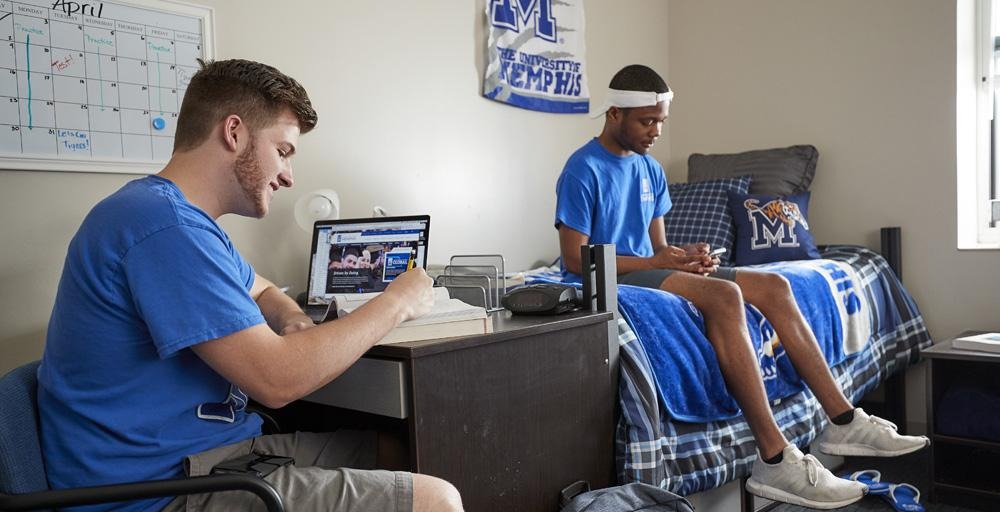 Two male students lounging in Living Learning Complex (LLC) double bed room with UofM blue and gray paraphernalia throughout; students wearing blue t-shirts, Tiger Athletics M-logo on the wall, calendar post on the wall. 