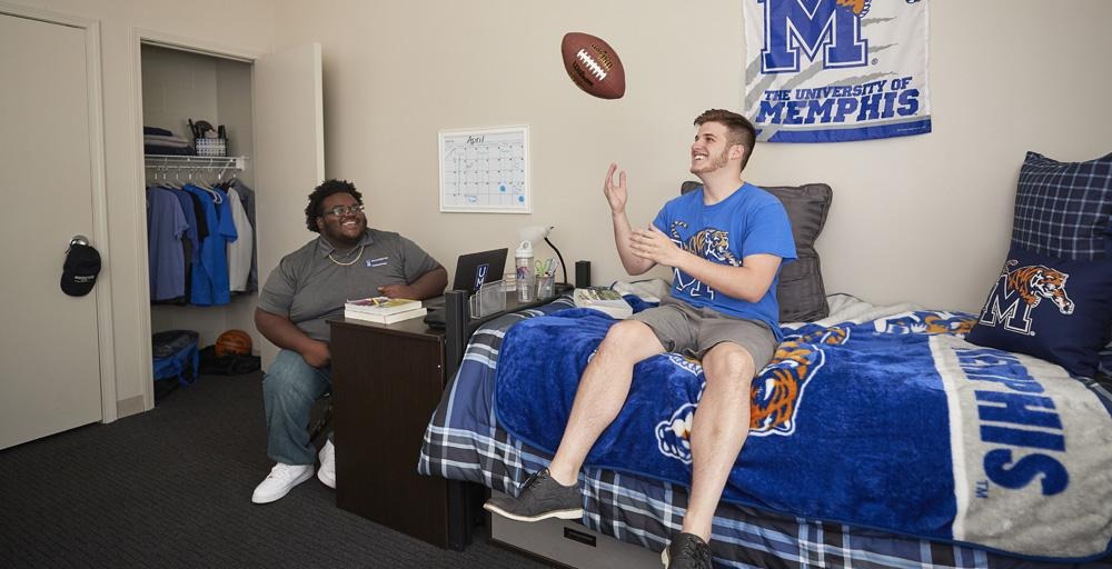Two students relaxing, one tossing football in the air sitting on a bed within Living Learning Complex with UofM blue and gray paraphernalia within the room; bed cover, signage on the wall, Tiger logo pillow. 