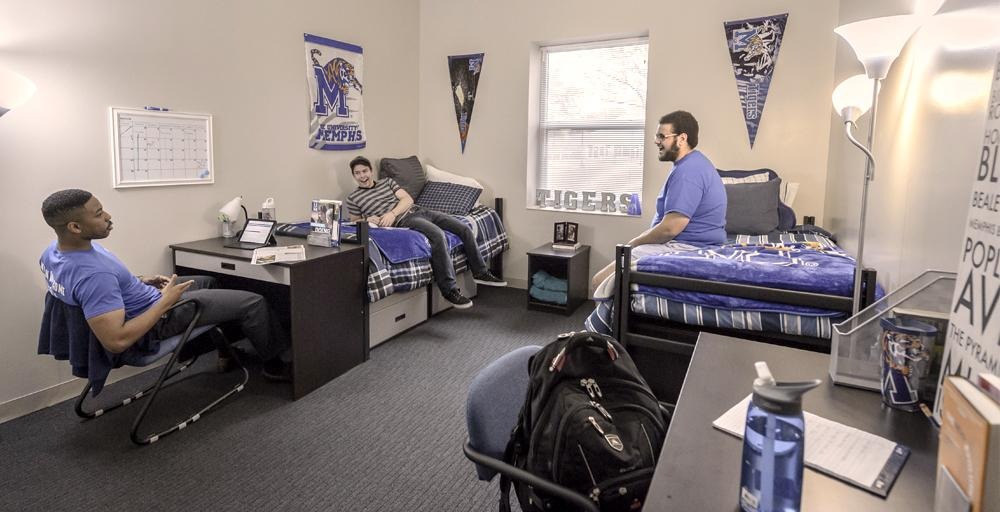 Three male students chatting, relaxing, two lounging on separate beds, one sitting with laptop at a desk in a Living Learning Complex double bedroom consisting of UofM blue and gray logo signage banners on the wall. 