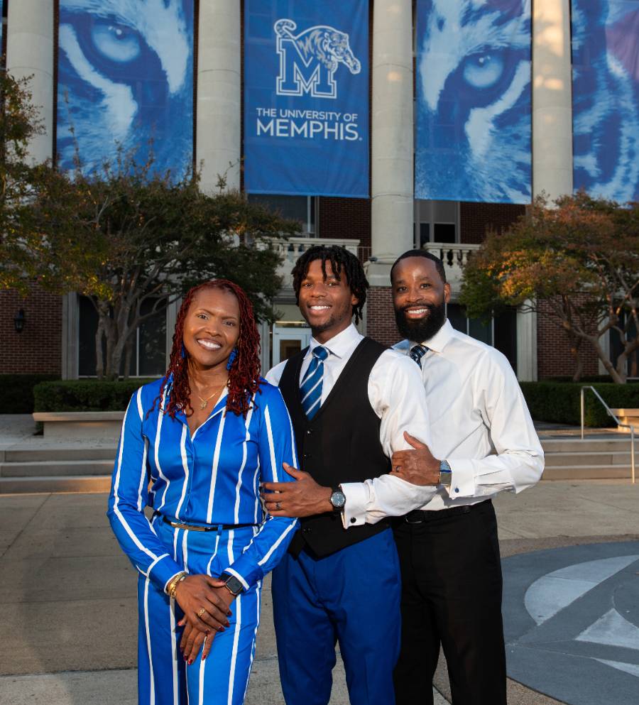 standing in front of the admin building, melody bradley in blue and white stripe outfit, flanked by son Chris Bailey in black vest and blue pants, and Andrew bailey, dad in button down white shirt. They are the 2025 UofM family of the year!