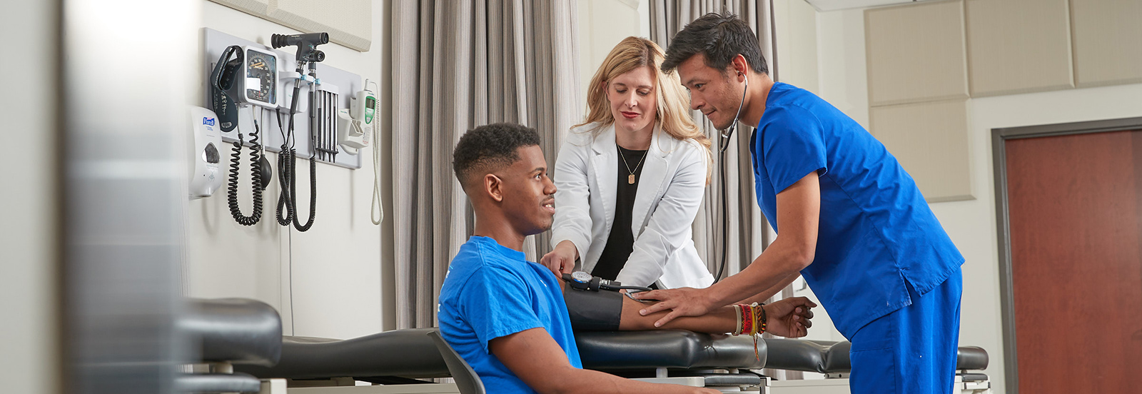 male medical student takes blood pressure of other male student while instructor in white lab coat offers guidance