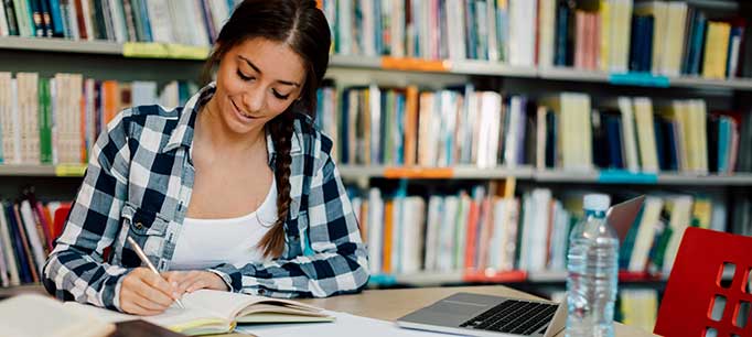 UofM Global Social taking notes in library while working on a laptop
