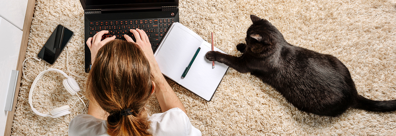 girl laying in floor typing on laptop while black cat plays reaches for her pencil laying next to her on a notebook