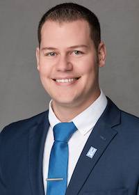 Professional headshot of a smiling individual wearing a dark blue suit, white dress shirt, and blue tie. A lapel pin with the letters 'U' and 'M' is visible on the jacket. The background is a plain gray, creating a formal and polished look.